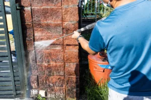 A person in a blue shirt pressure washes a brick wall with a high-pressure water spray.