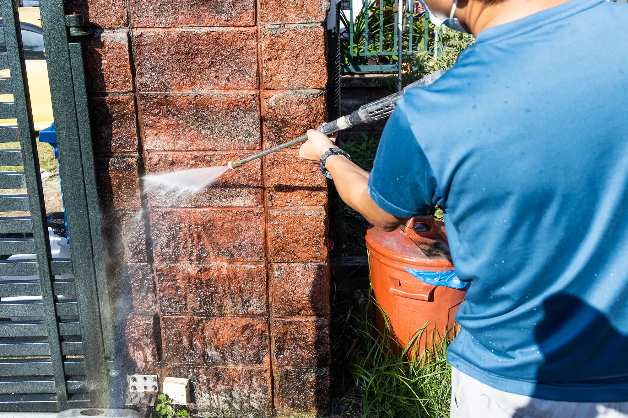 A person in a blue shirt pressure washes a brick wall with a high-pressure water spray.